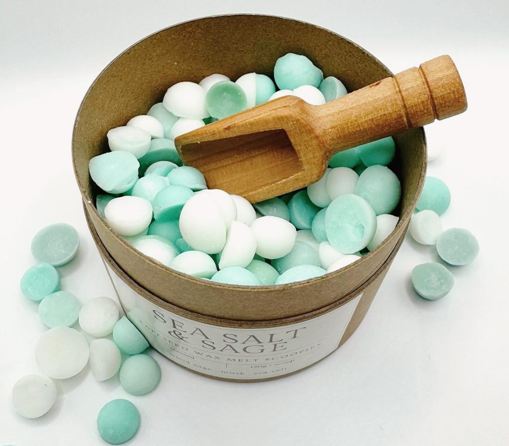 A kraft cardboard tub with wooden scoop containing green and white wax melt drops.